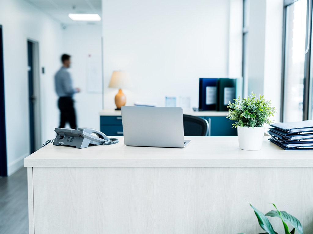 A clean front desk with a laptop, a desk phone and a small plant, softly lit by window light, with a blurred figure in t